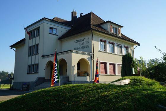 Eingang des Schulhauses mit Fahne und Hügel im Vordergrund