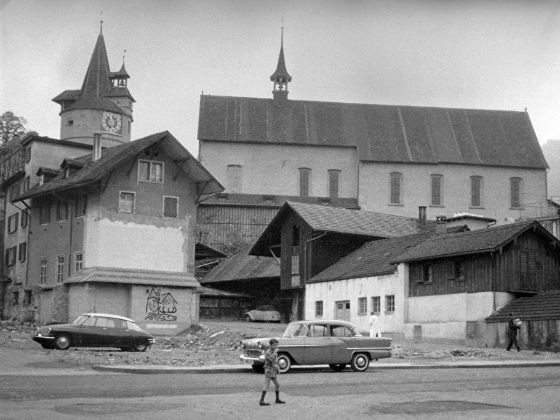 Schwarz-weisse Darstellung des Kapuzinerklosters in Zug mit Kapuzinerturm und Friedensglocke im Jahr um 1960. Im Vordergrund sind zwei Autos aus dieser Zeit sichtbar und ein Junge in Kniesocken.