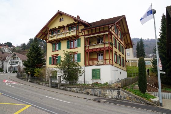 Strasse im Vordergrund, gelbes Chalet mit Garten im Vordergrund und Balkon